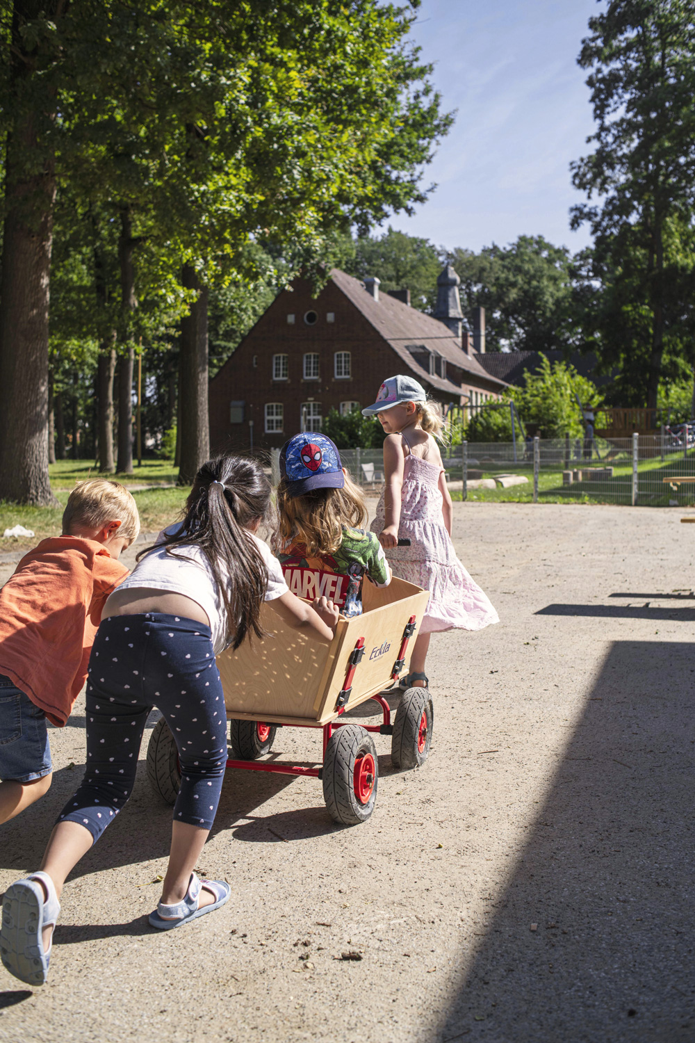 Drei Kinder schieben einen Bollerwagen mit einem Kind über einen Weg