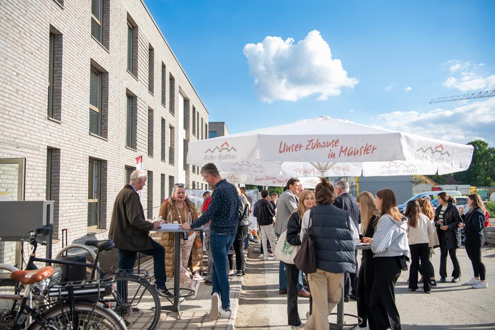 Eine Gruppe Menschen steht unter einem Sonnenschirm. Links ein Neubau.